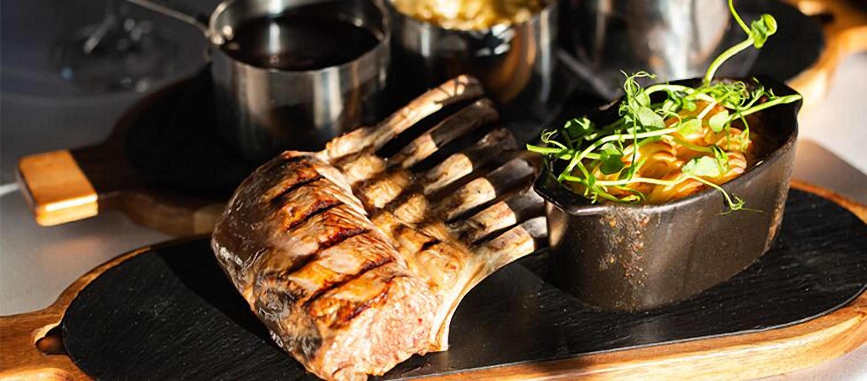 A grilled lamb chop on a wooden board with a bowl of greens and a pot in the background.