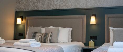 A hotel room with two beds, each with a headboard, pillows, and a blanket. There are wall-mounted lamps on either side of the beds.