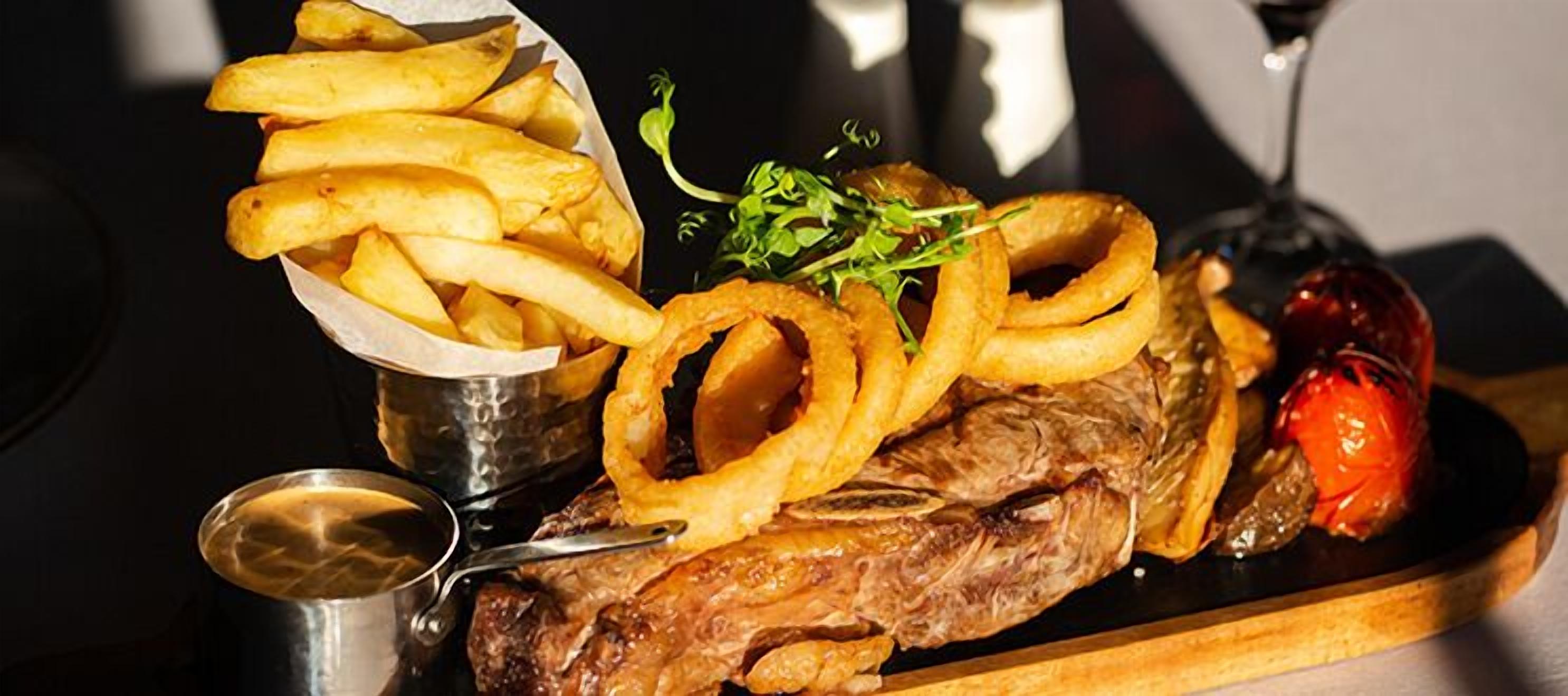 A steak with onion rings, a side of fries, and a small bowl of sauce.