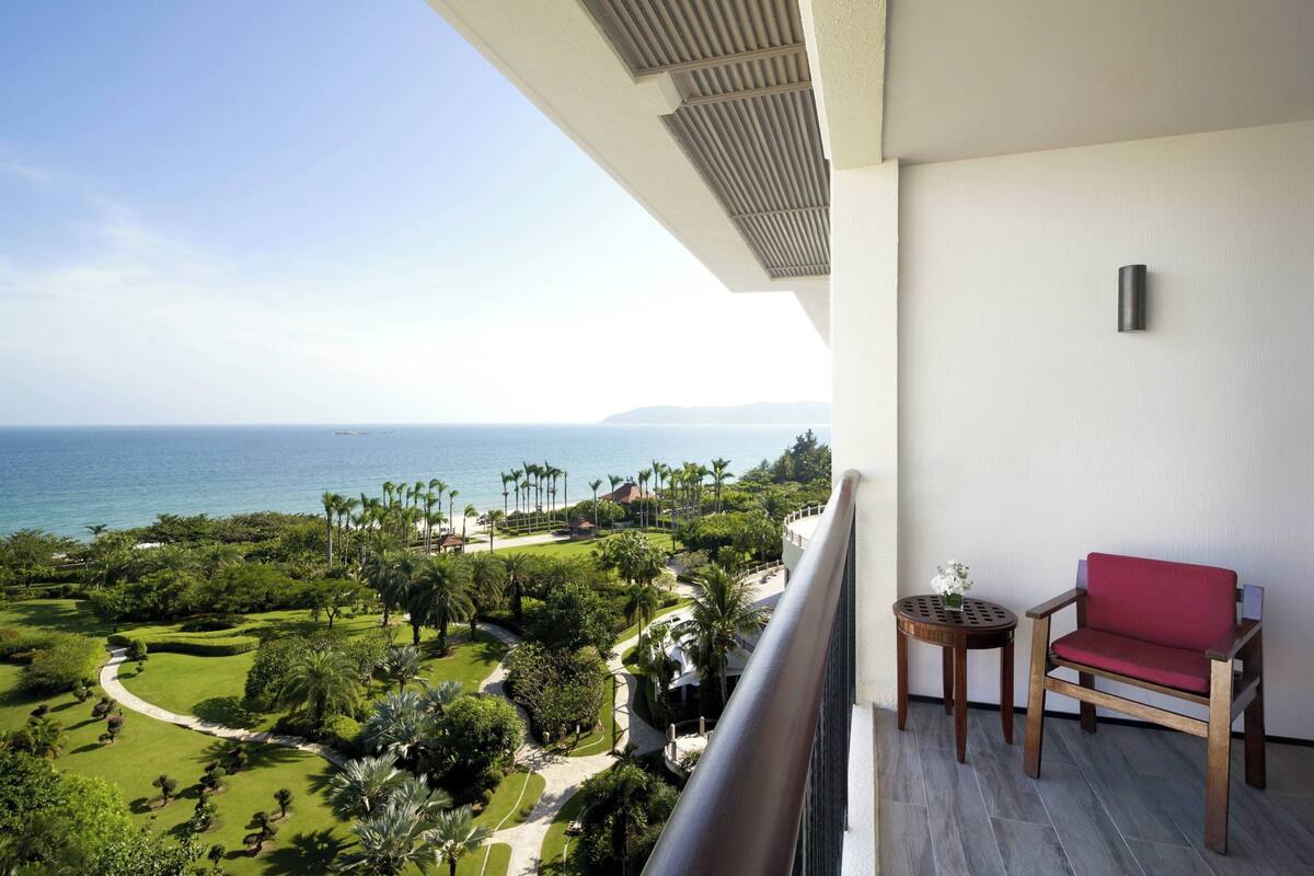 Un balcon avec une chaise rouge et une petite table, donnant sur un jardin bien entretenu et l’océan.