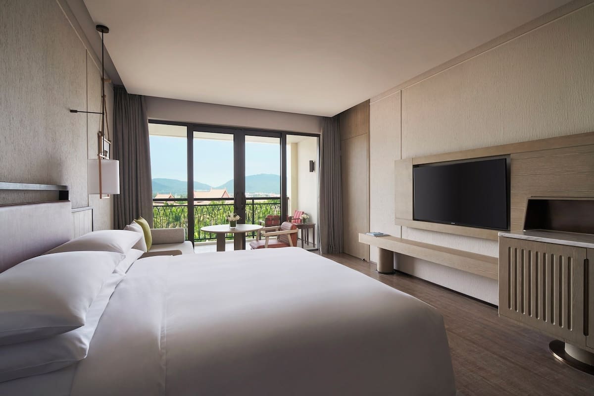 Une chambre d’hôtel avec un grand lit, une télévision, un balcon avec une table et des chaises, et une vue sur les montagnes.