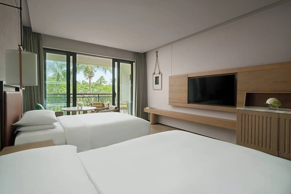 Une chambre d’hôtel moderne dotée d’un grand lit, d’un téléviseur fixé au mur et d’un balcon offrant une vue sur les palmiers.