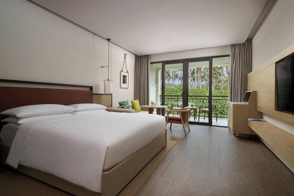 Une chambre d’hôtel avec un grand lit, un bureau, une chaise, une télévision et un balcon donnant sur un espace verdoyant.