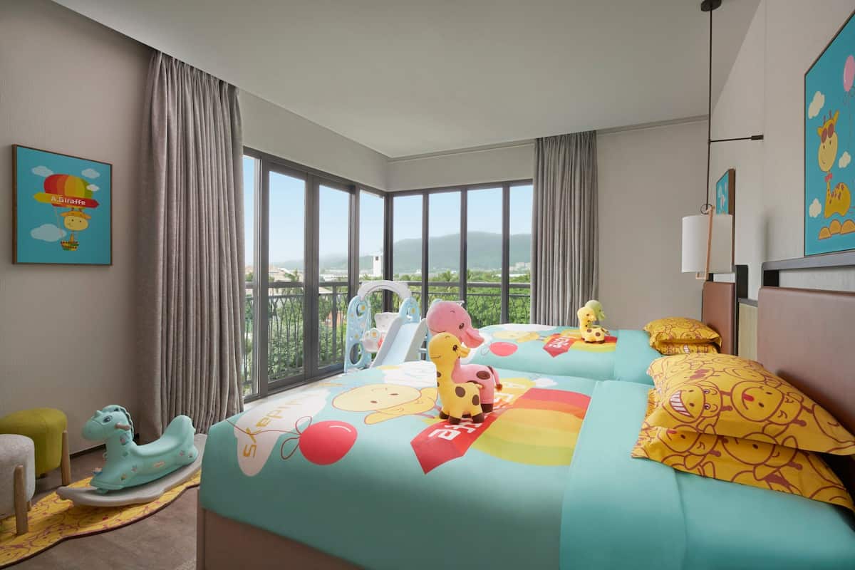 Une chambre d’hôtel avec une grande fenêtre, un lit avec une literie aux couleurs vives, un cheval à bascule pour enfant et un petit espace de jeux.