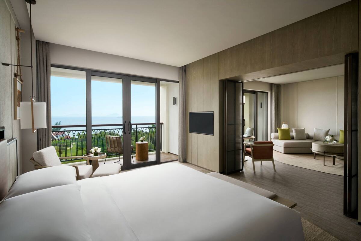 Une chambre d’hôtel moderne dotée d’un grand lit, d’un balcon avec vue, d’un coin salon avec un canapé et des fauteuils, et d’un téléviseur à écran plat.