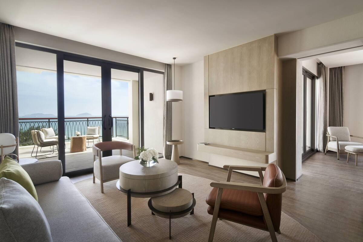 Une chambre d’hôtel moderne dotée d’un grand balcon, d’une table basse ronde, d’un canapé et d’un téléviseur à écran plat fixé au mur.