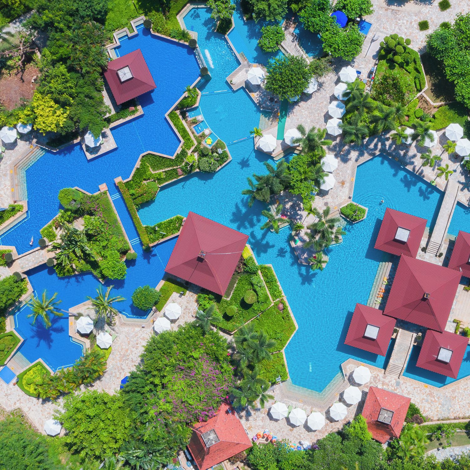 A large, multi-pool resort with separate sections for each pool, surrounded by lush greenery and red-roofed structures.