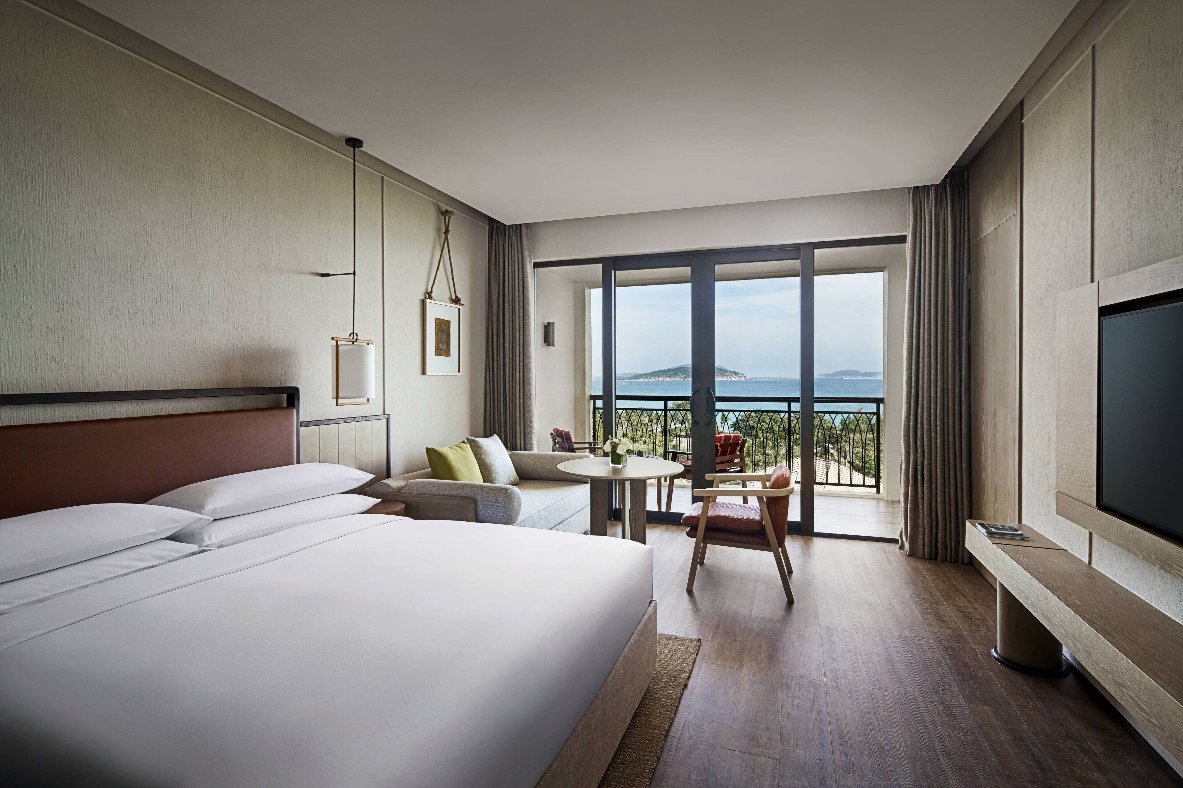 Habitación de hotel con una cama grande, televisión, balcón con vista al mar y zona de comedor con mesa y sillas.