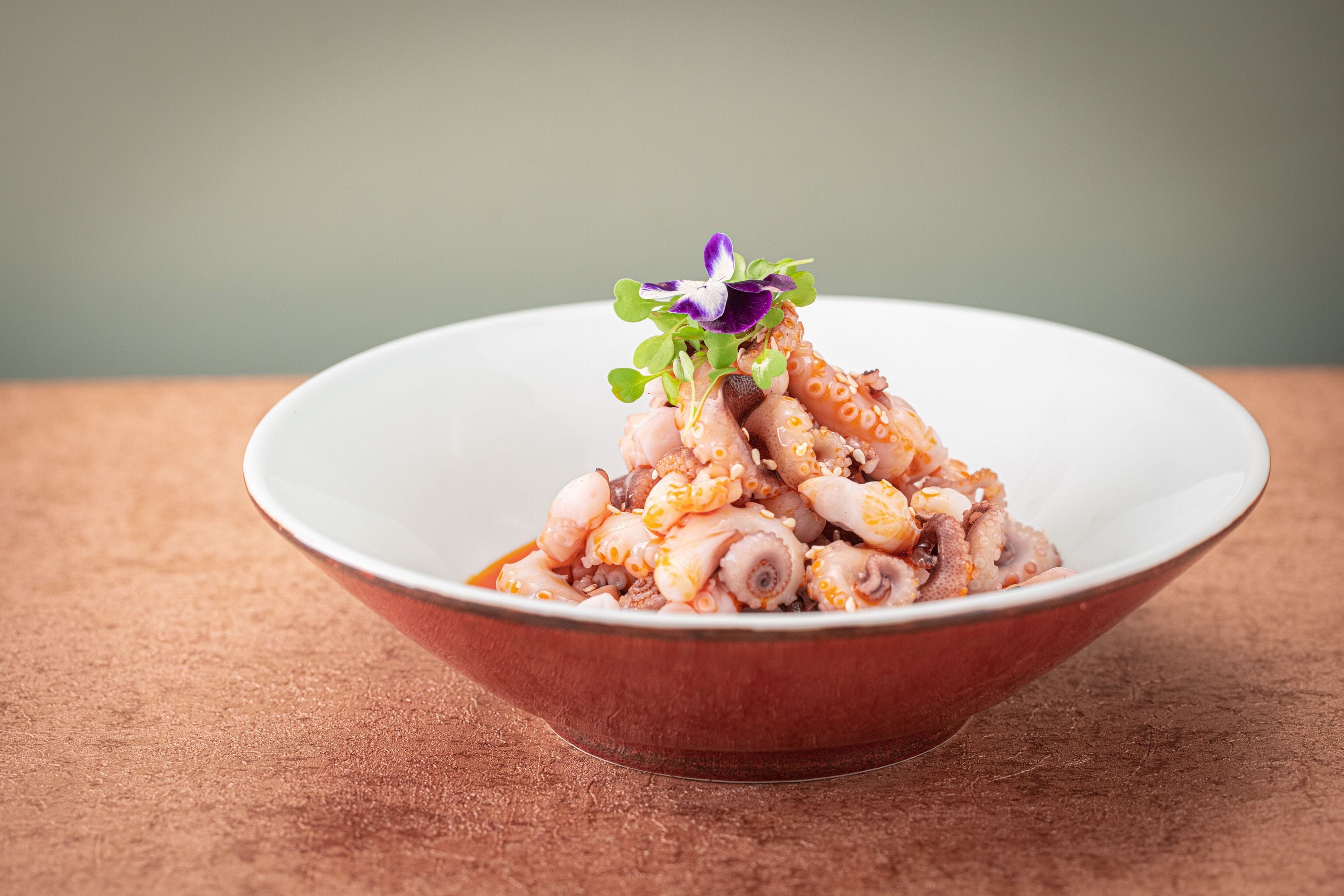 A bowl of octopus with a purple flower garnish on a textured surface.