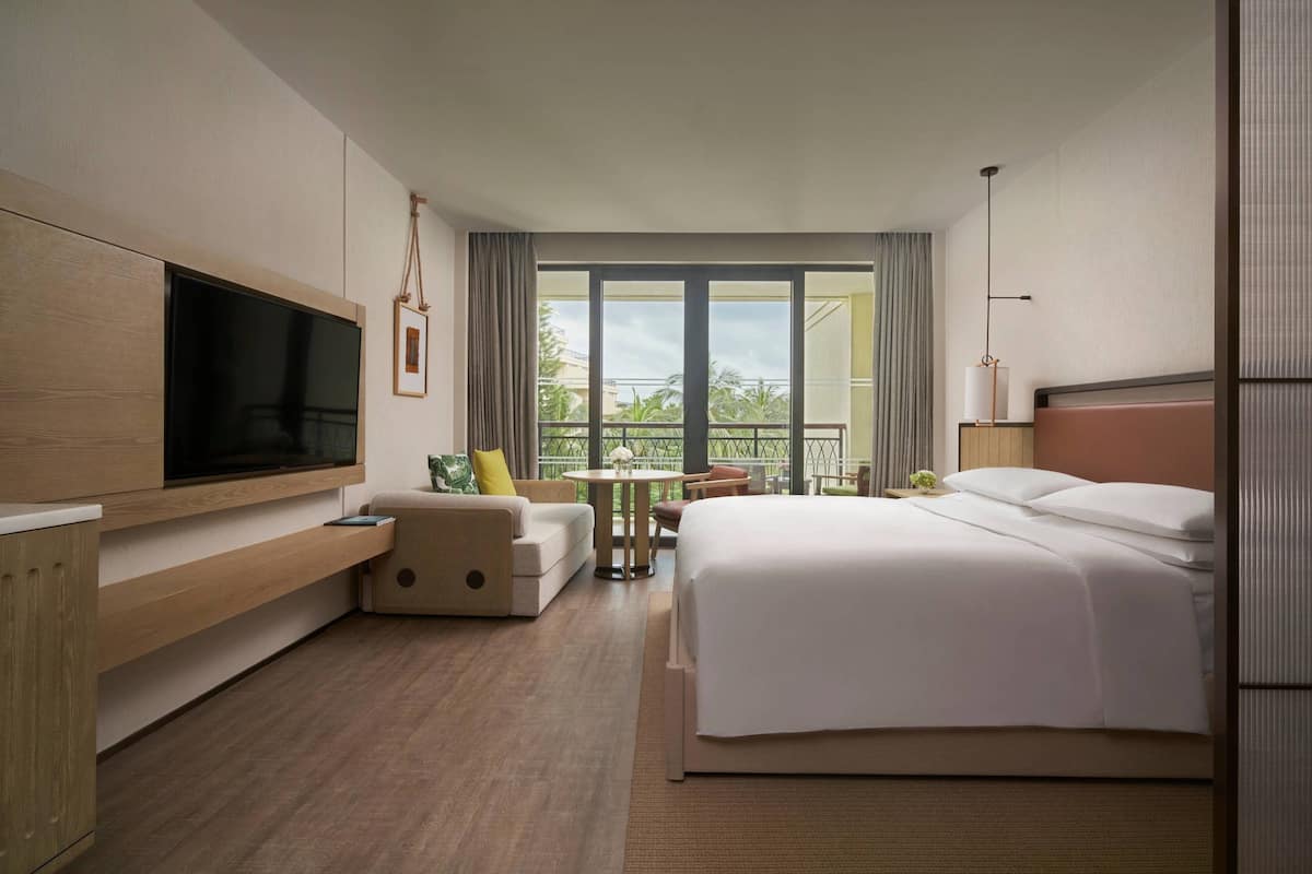 Une chambre d’hôtel dotée d’un grand lit, d’un téléviseur fixé au mur, d’un coin salon avec une table et d’un balcon offrant une vue sur la verdure.