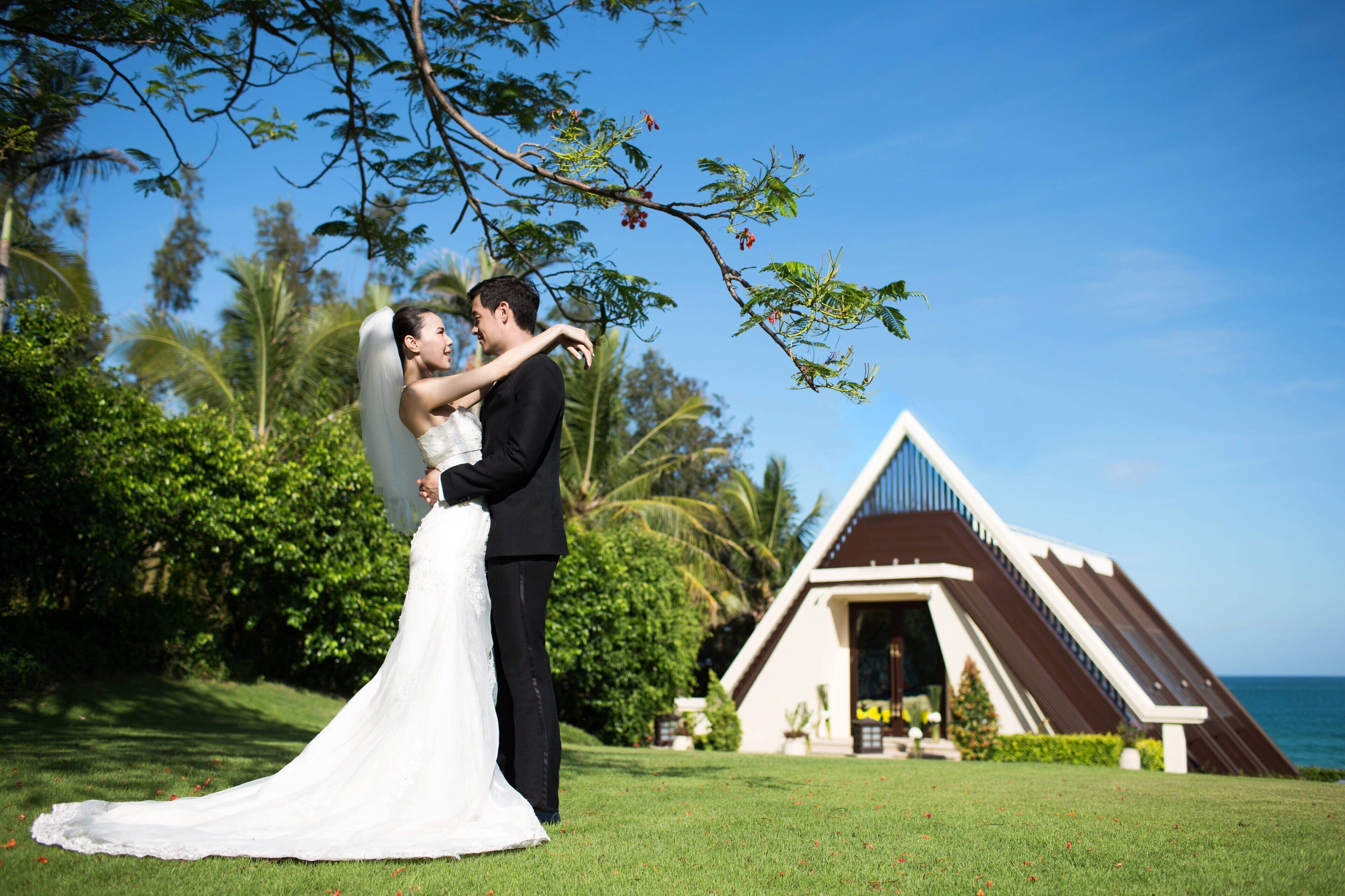 A couple embracing in a lush outdoor setting with a building featuring a triangular roof and a view of the ocean.