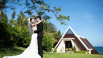 A couple embracing in a lush outdoor setting with a building featuring a triangular roof and a view of the ocean.
