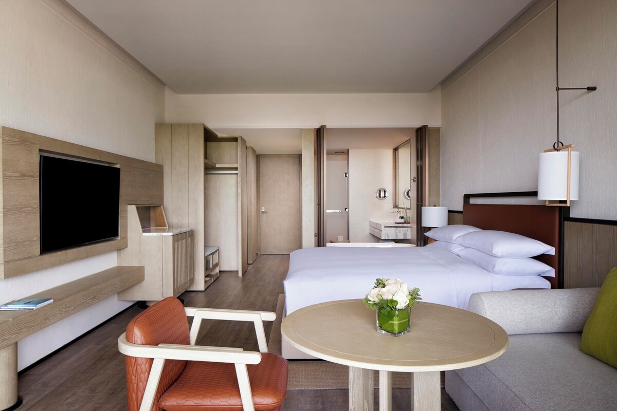 Quarto de hotel com cama, televisão, área de estar com mesa e banheiro visível através de uma porta aberta.
