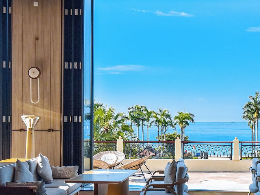 A modern interior with a large window overlooking a beach, furnished with a sofa, chairs, and a table.