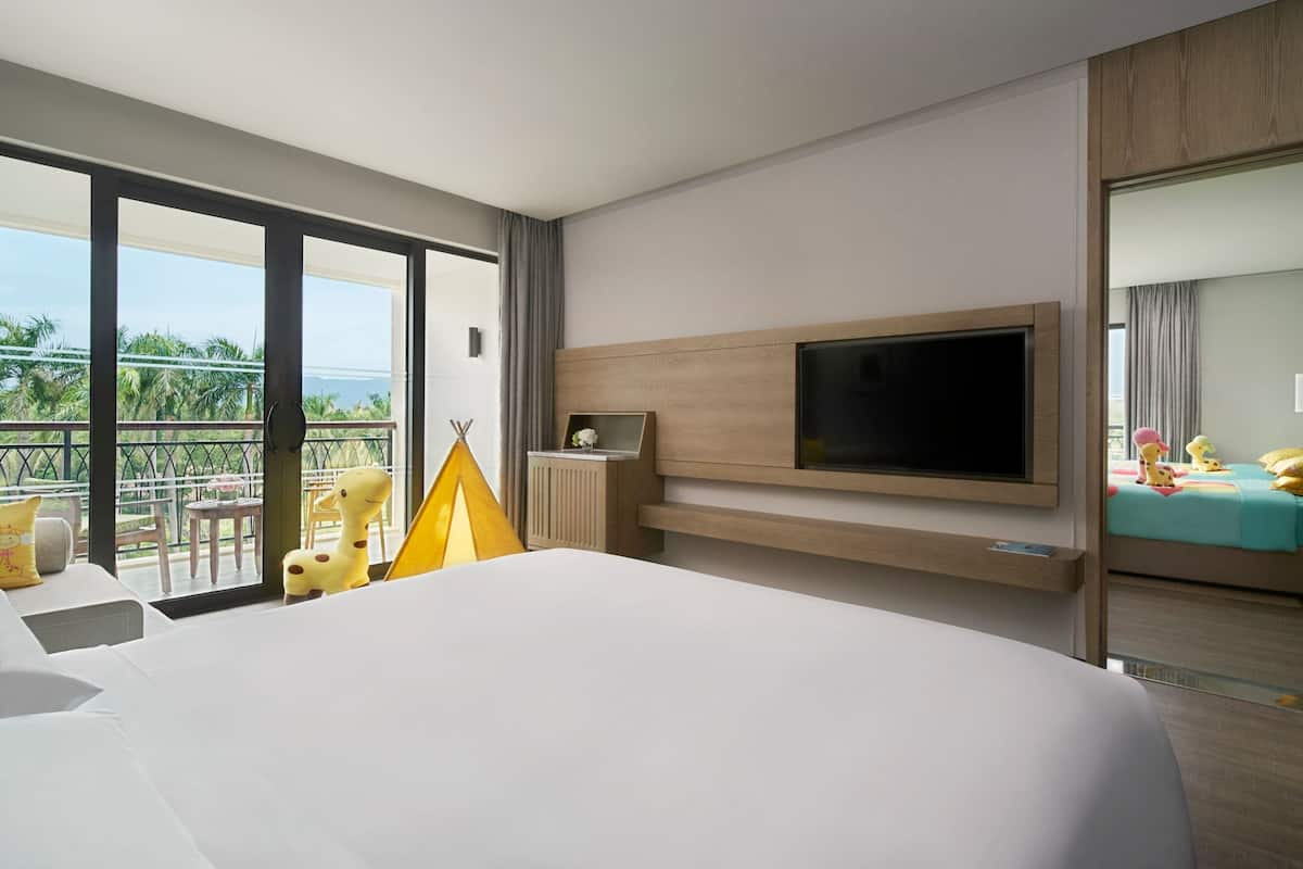 Une chambre d’hôtel avec un grand lit, un téléviseur fixé au mur, un balcon avec vue sur la mer et une petite table avec une lampe.