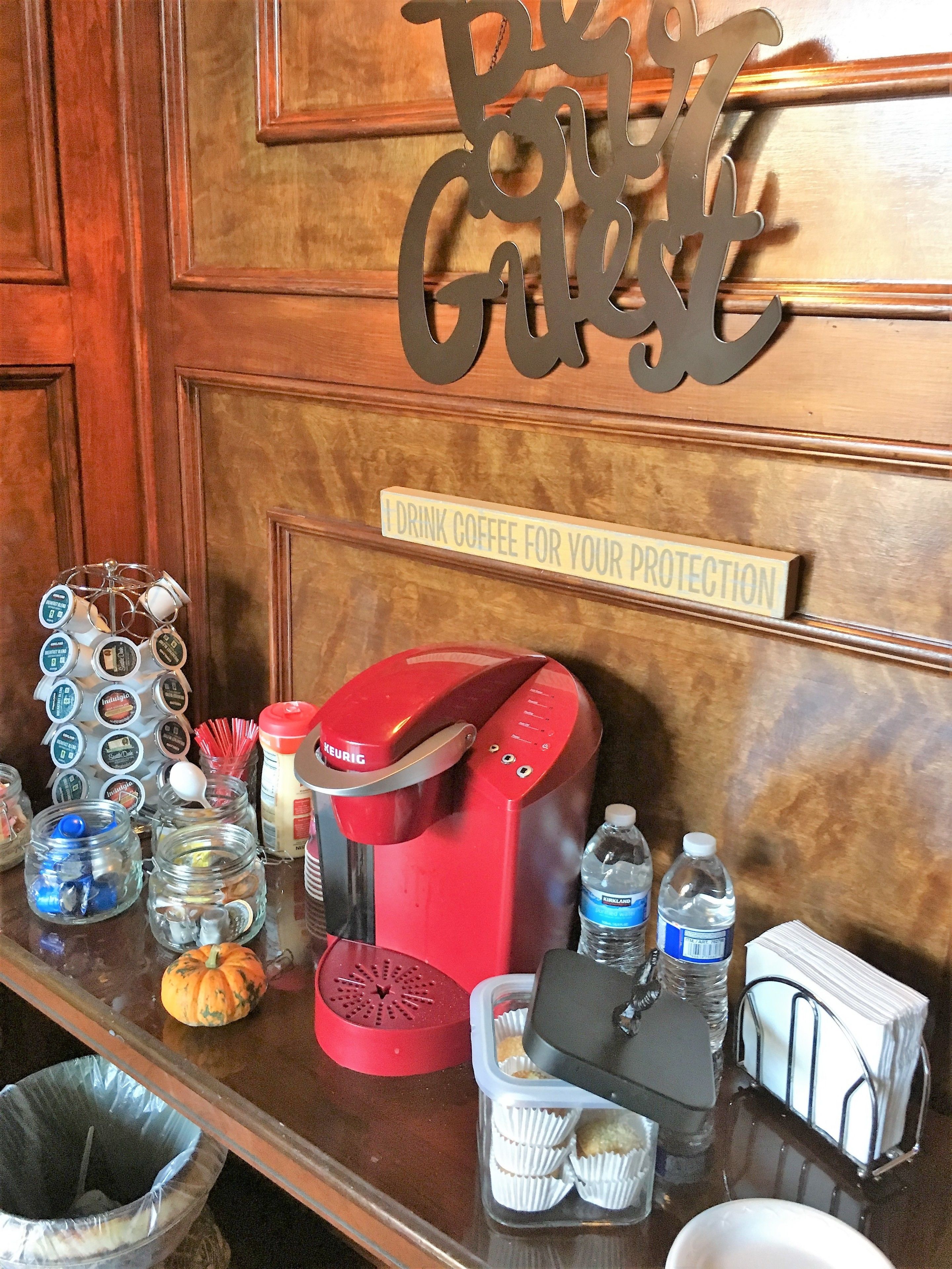 A coffee station with a Keurig machine, coffee pods, a water dispenser, and a sign that reads "I drink coffee for your protection."