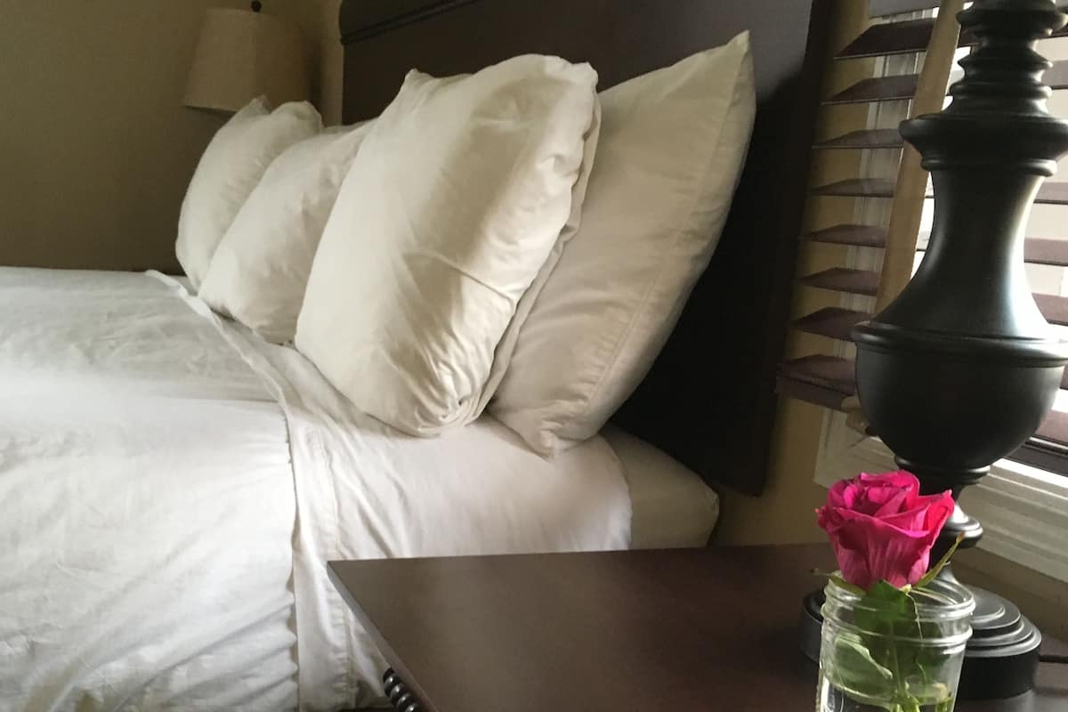 A neatly made bed with white linens and pillows, a dark wooden nightstand with a vase containing a single pink rose, and a lamp with a beige shade.