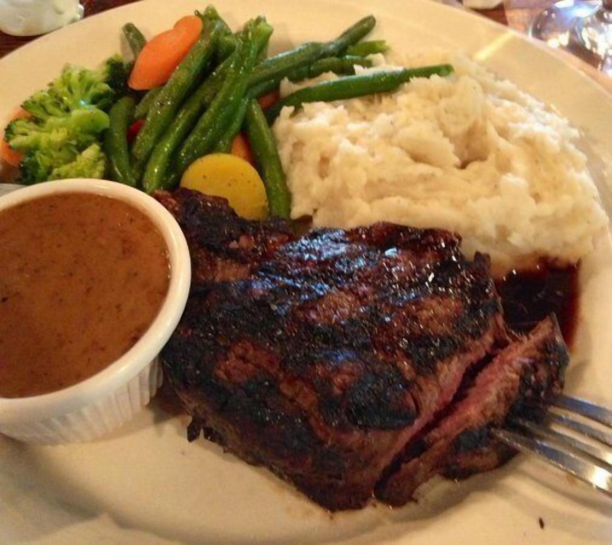 A plate with a steak, mashed potatoes, green beans, carrots, and a gravy bowl.