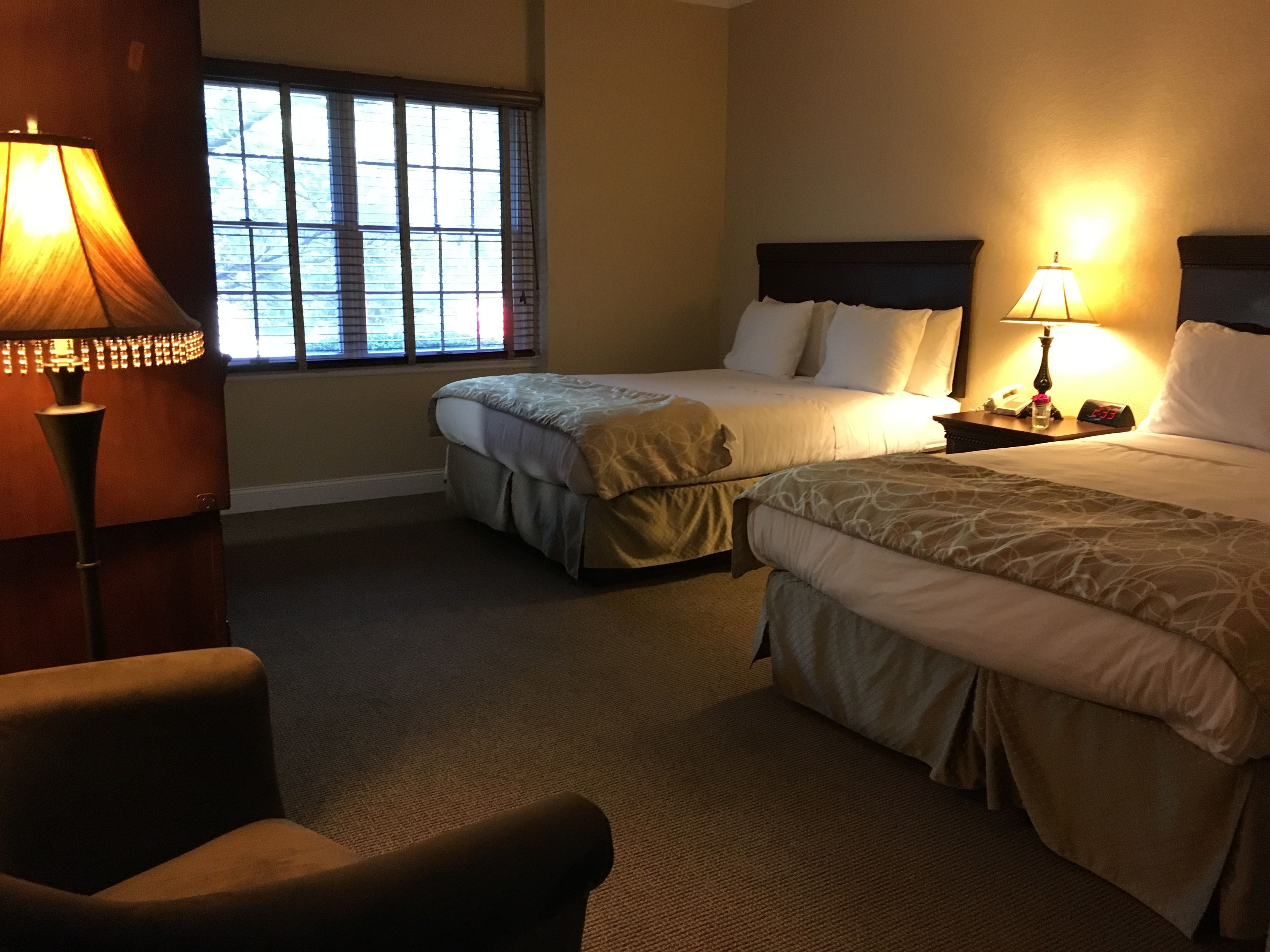 A hotel room with two beds, a chair, a nightstand with a lamp, and a window with blinds.
