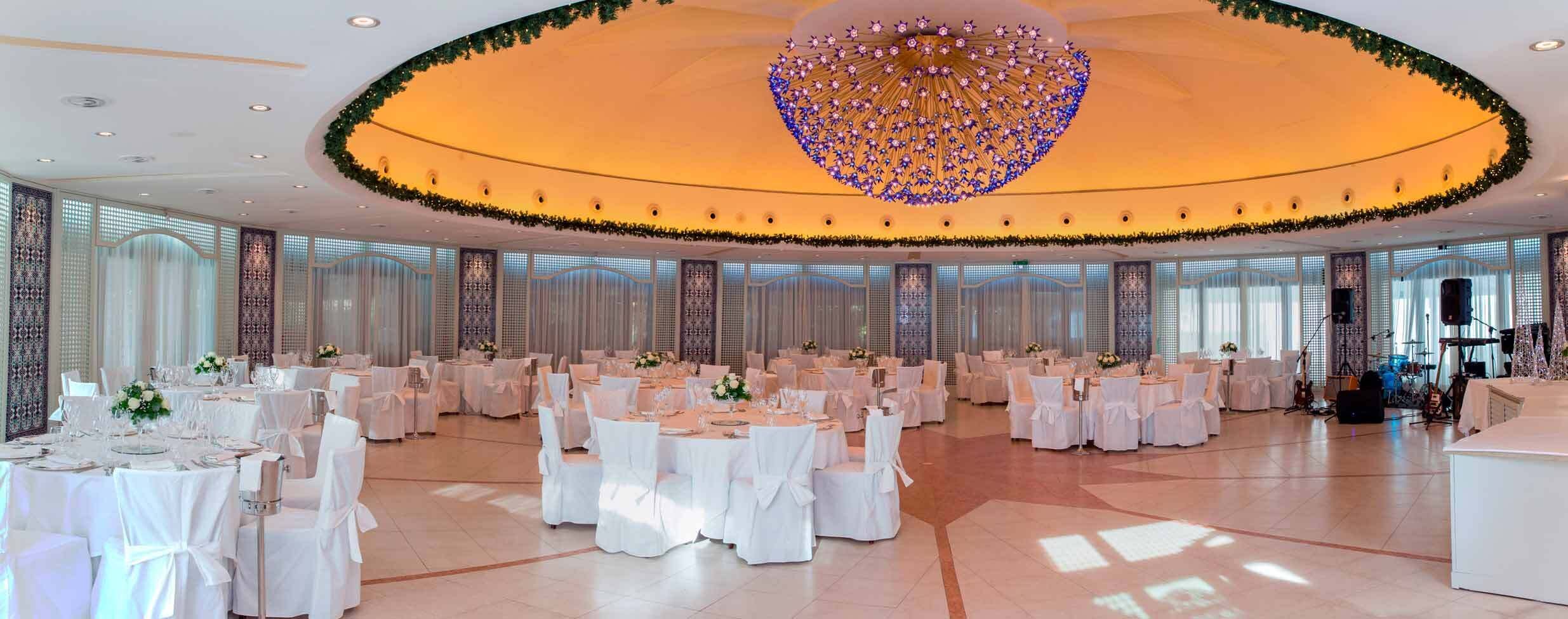 A spacious banquet hall with round tables, white tablecloths, and a large decorative chandelier.
