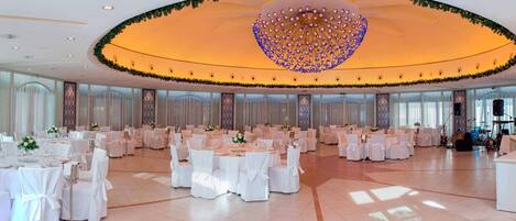 A spacious banquet hall with round tables, white tablecloths, and a large decorative chandelier.