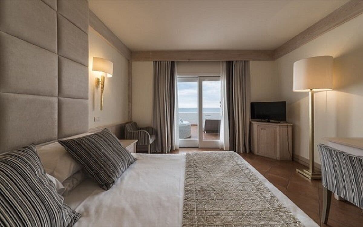 Et hotelværelse med en stor seng, et fjernsyn, et skrivebord og havudsigt gennem en glasdør.