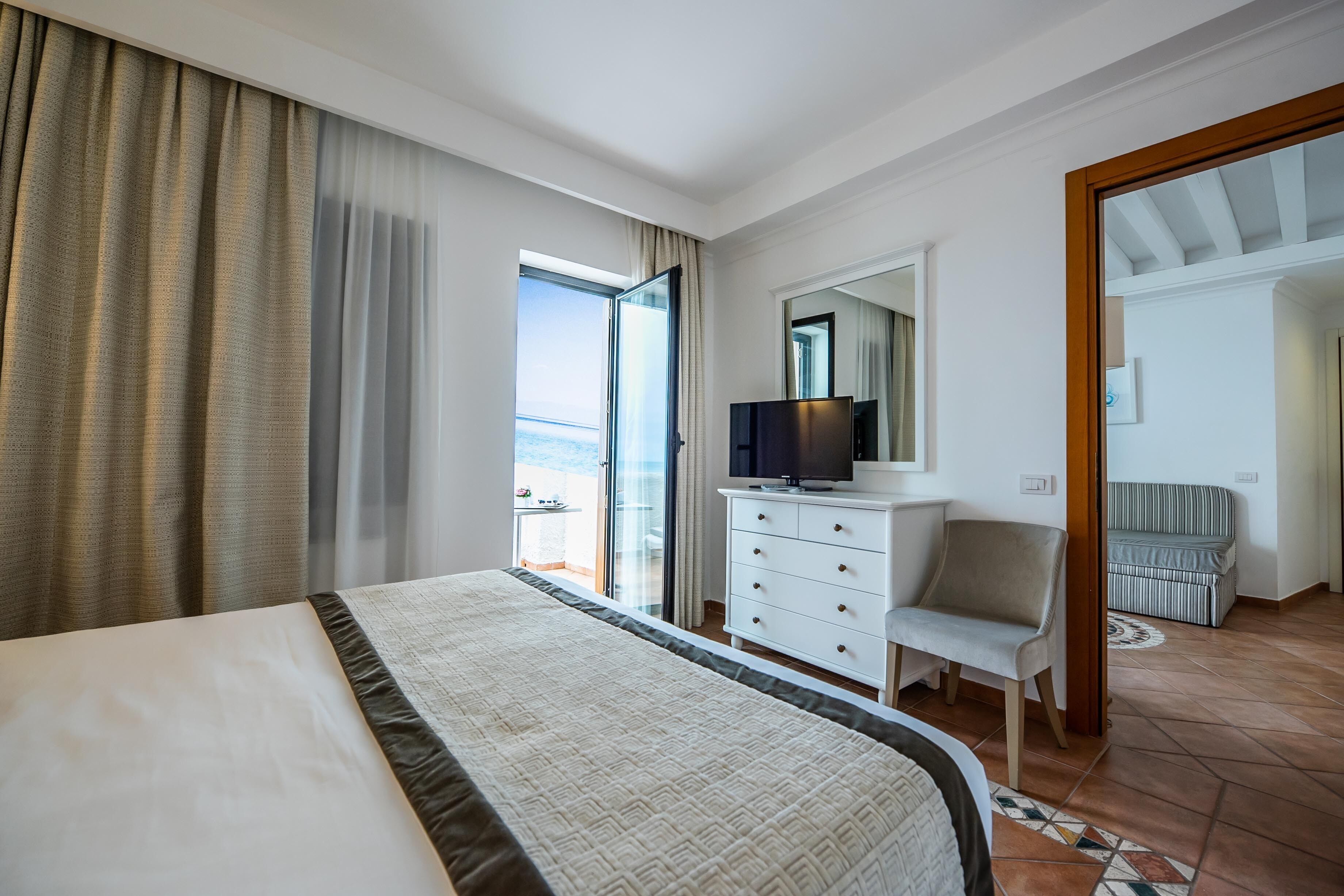Una camera d'albergo con un letto, una televisione, una sedia e vista mare attraverso un'ampia finestra.