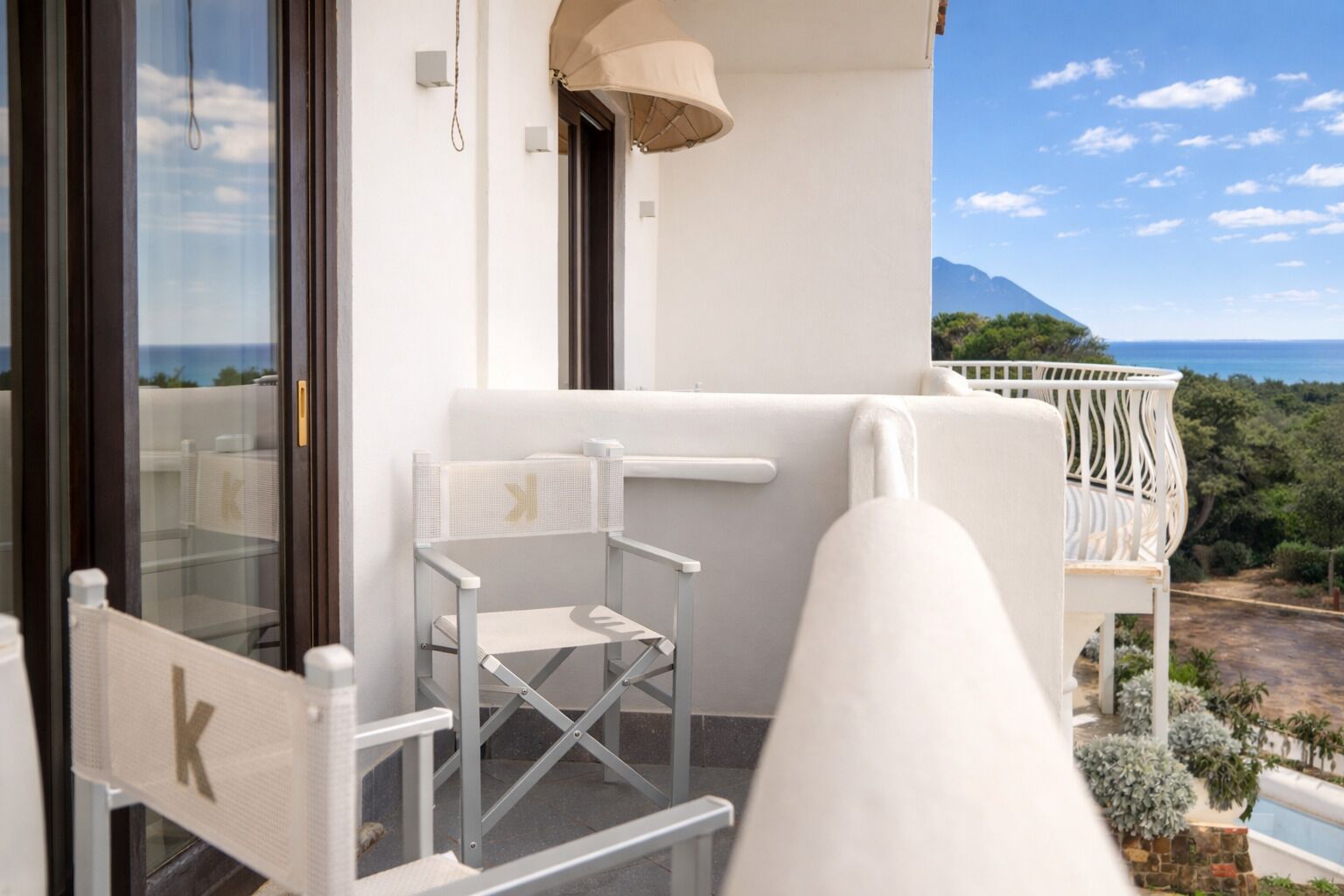 Un balcón con una silla blanca, una mesa pequeña y vistas al mar y a las montañas.
