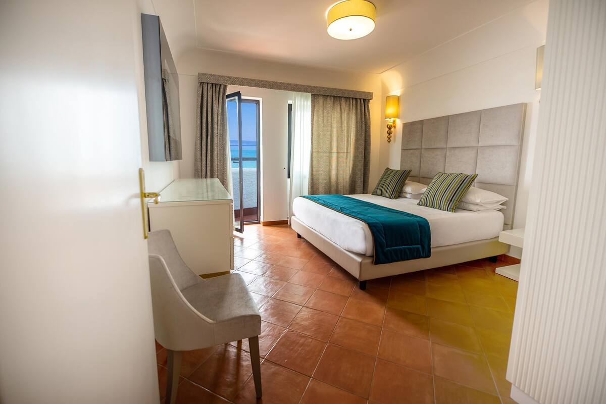 Une chambre d’hôtel avec un lit, un bureau, une chaise et une vue sur la mer, visible par la porte.
