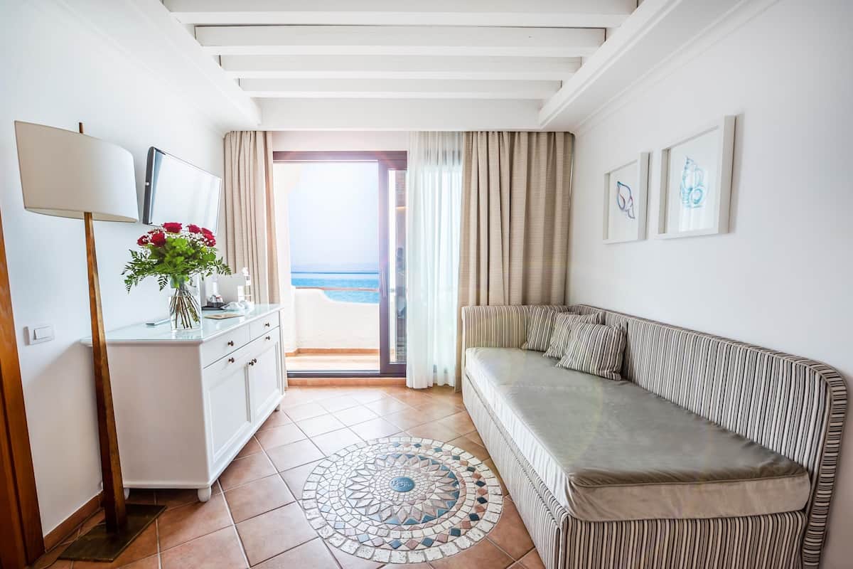 Une pièce comprenant un canapé, un bureau avec une lampe, un vase de fleurs et une vue sur la mer, visible à travers une porte coulissante.