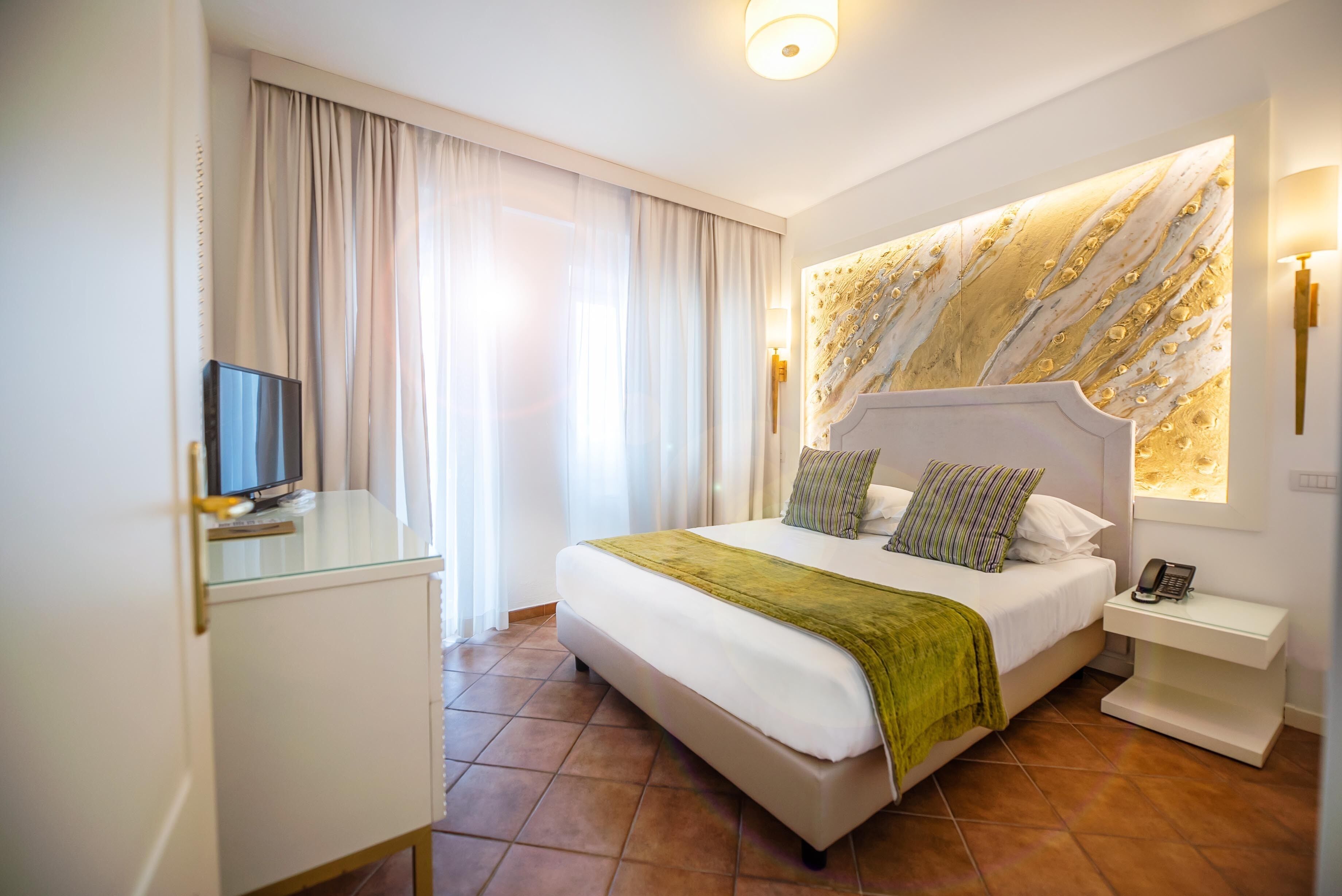 Habitación de hotel con una cama grande, un televisor sobre un mueble de cristal, una mesita de noche con un teléfono y un panel decorativo en la pared.