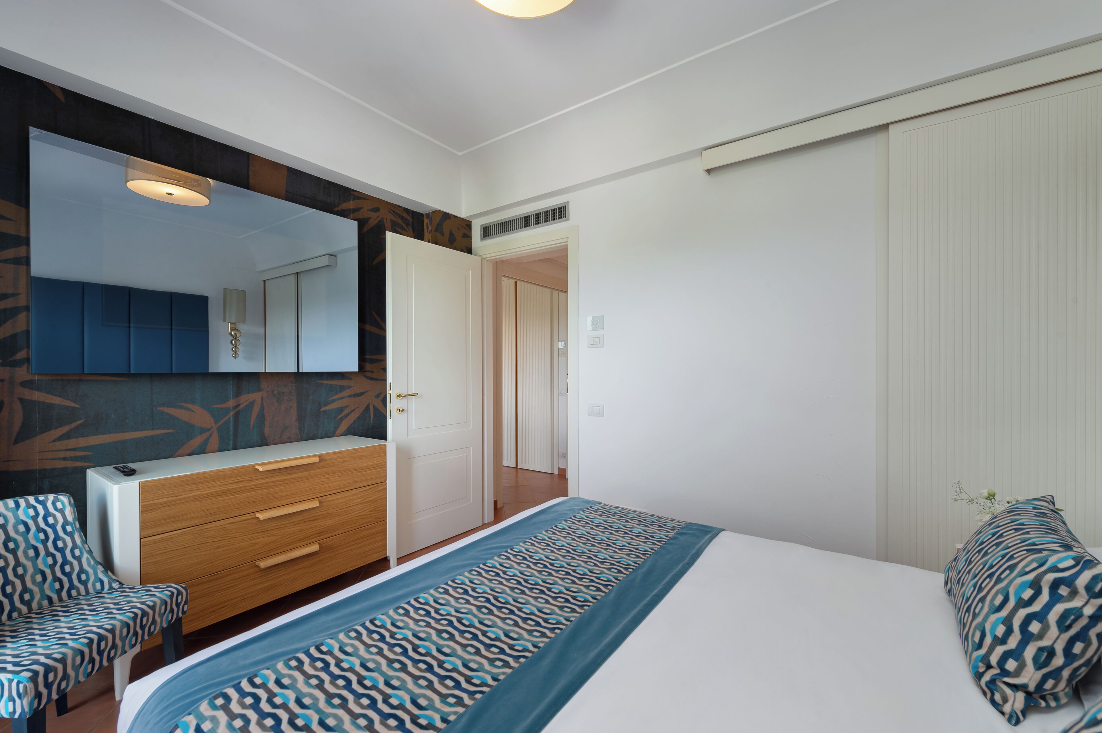 A modern bedroom with a large bed, a dresser, a mirror, and a door leading to another room.