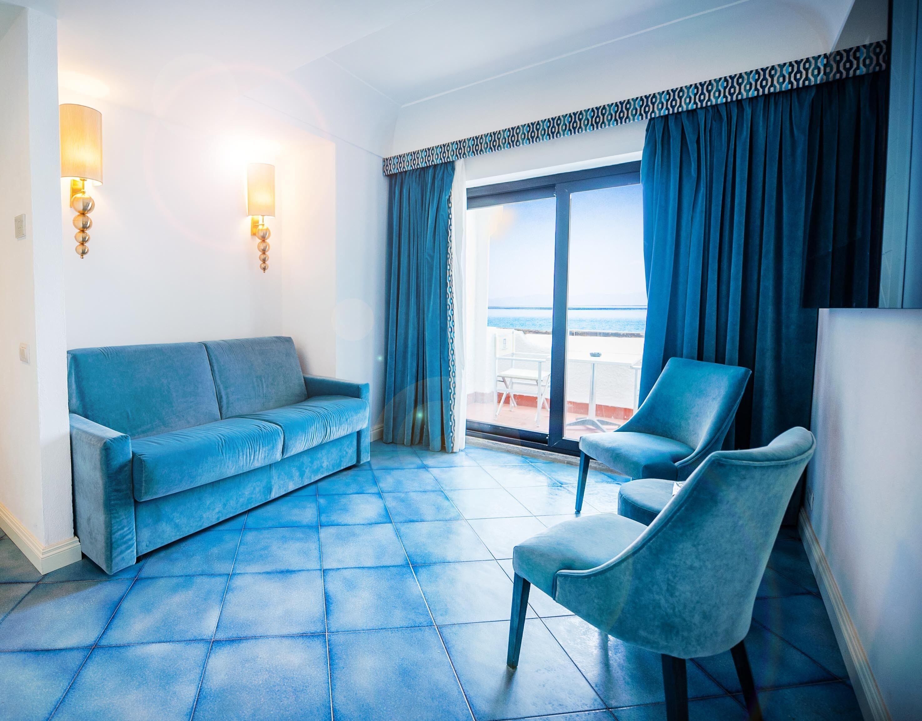 A room with a blue sofa, a blue armchair, and a large window with a view of the sea.