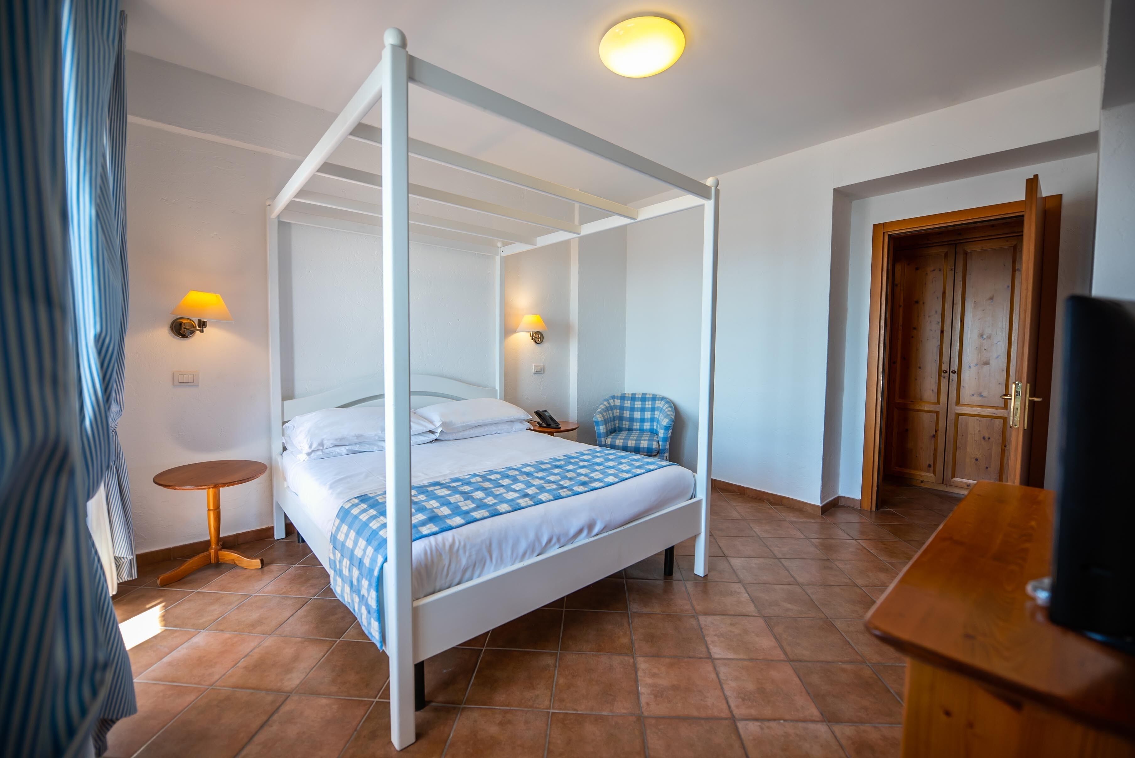 A four-poster bed with a checkered bedspread, two bedside lamps, a small round table, and a wooden desk.