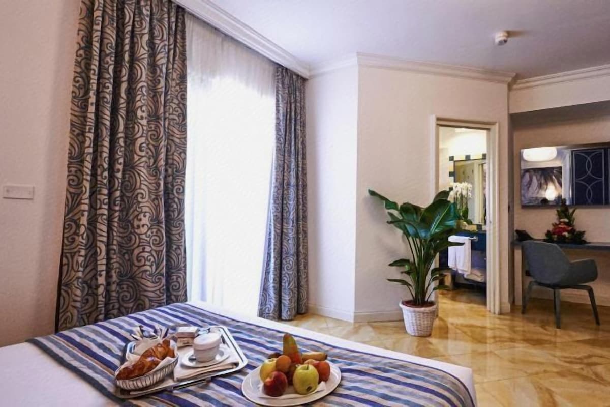 A hotel room with a bed, breakfast tray, potted plant, desk, chair, and a view of the hallway.
