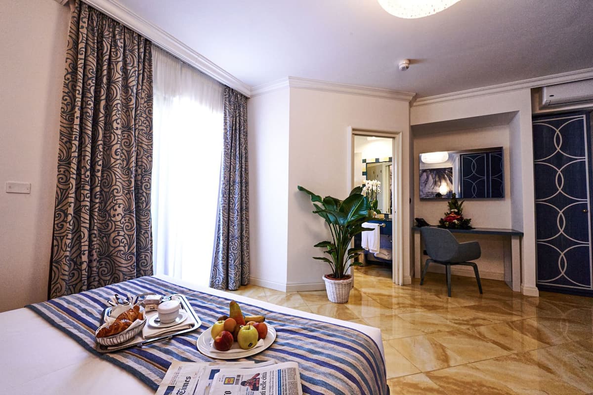 Une chambre d’hôtel avec un lit, un plateau de petit-déjeuner, une assiette de fruits, une plante en pot, un bureau, une chaise et une salle de bain visible à travers une porte ouverte.