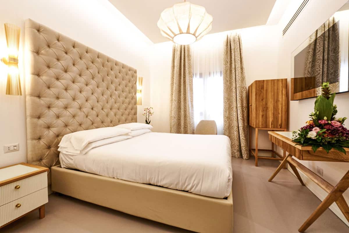 Une chambre d’hôtel avec une tête de lit capitonnée, une table de chevet en bois, un bureau agrémenté d’un vase de fleurs et un miroir.