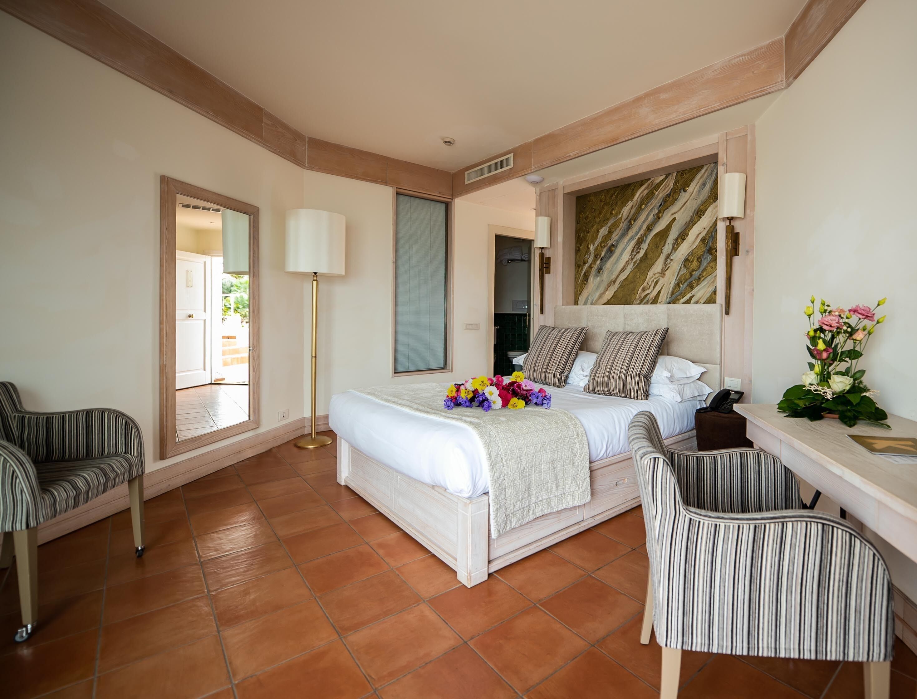 A hotel room with a bed, two chairs, a desk, a lamp, and a vase of flowers.