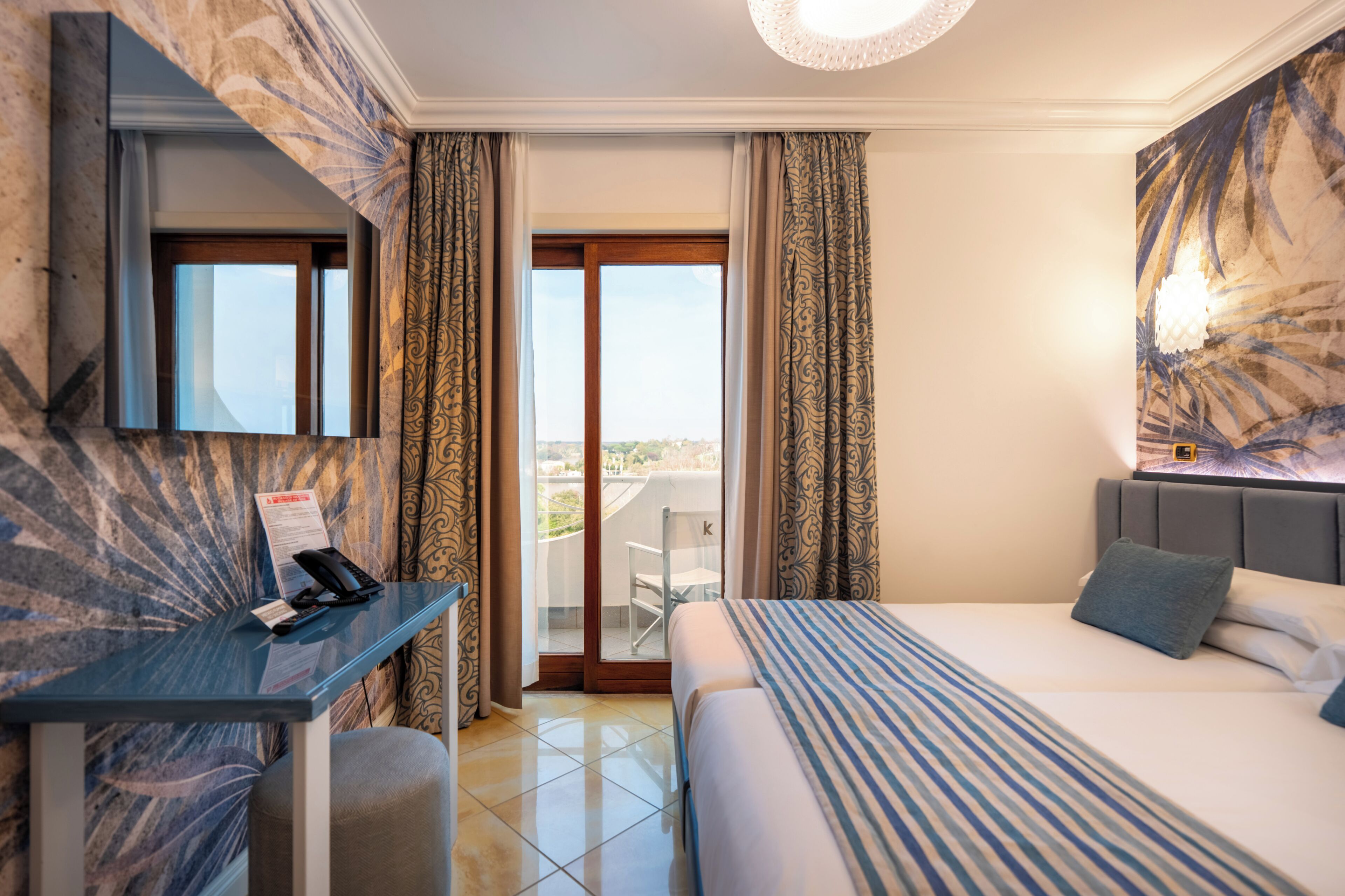Habitación de hotel con cama, escritorio con teléfono, una silla y un ventanal con cortinas.