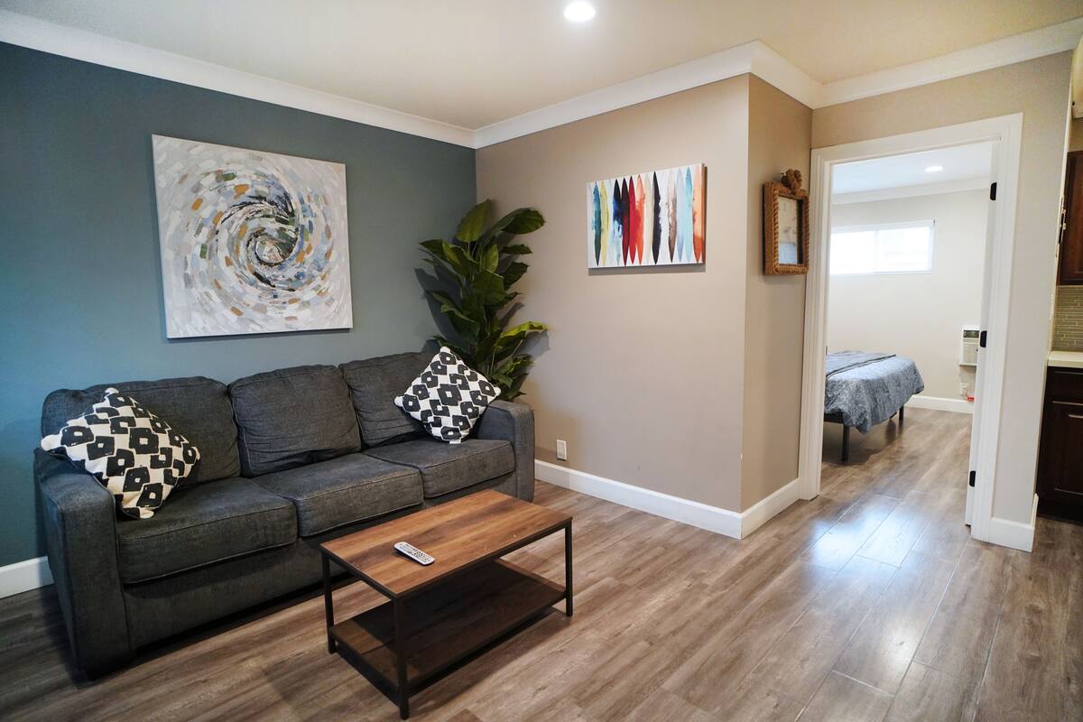 A living room with a gray sofa, a coffee table, and a large plant. There is a painting on the wall and a door leading to another room.