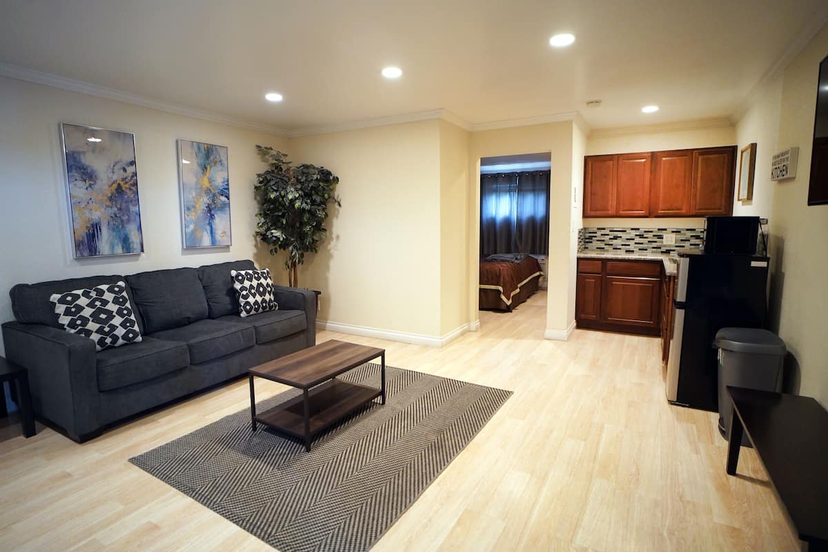A modern living room with a sofa, coffee table, and a rug. There is a kitchen area with wooden cabinets and a refrigerator.