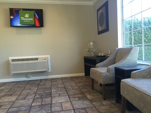 A room with a television displaying Panera Bread, a coffee maker, two armchairs, a radiator, and a window with a view of greenery.