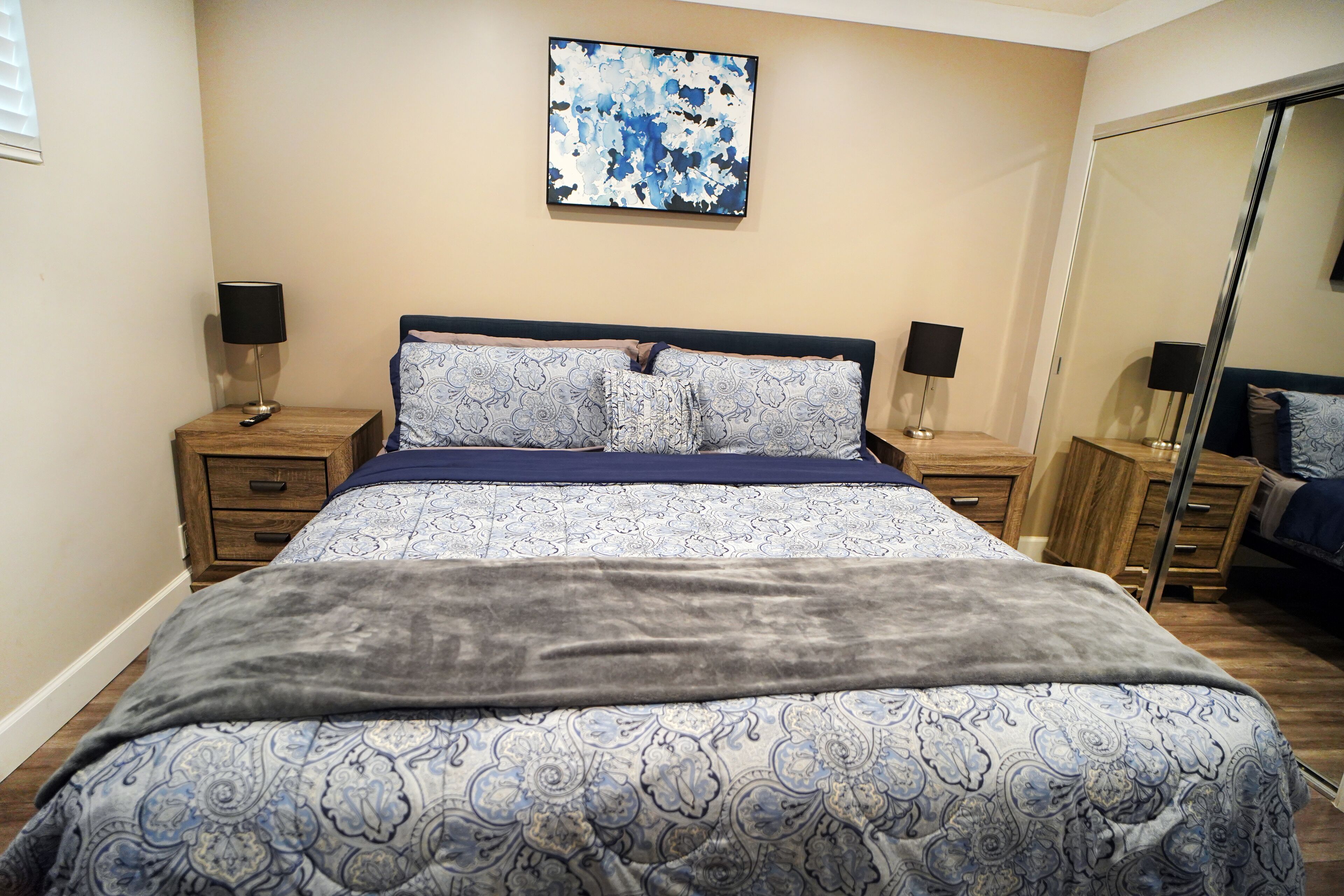 A bedroom with a bed, two bedside tables with lamps, a mirror, and a blue abstract painting on the wall.