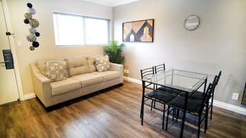 A living room with a beige sofa, patterned cushions, a glass-top dining table with chairs, a potted plant, and a wall clock.