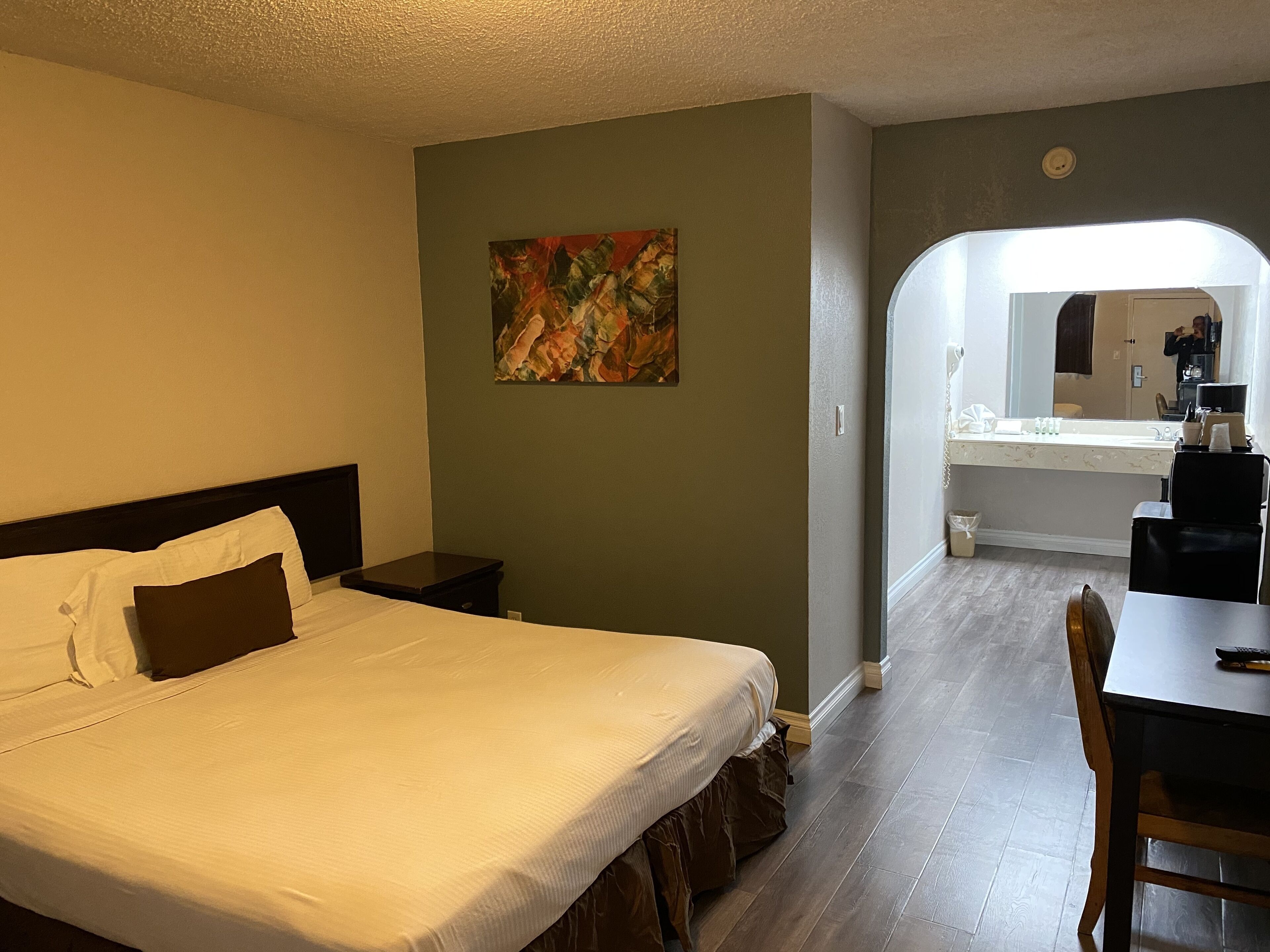 A hotel room with a bed, a small bedside table, a painting on the wall, and a view of the bathroom through an archway.