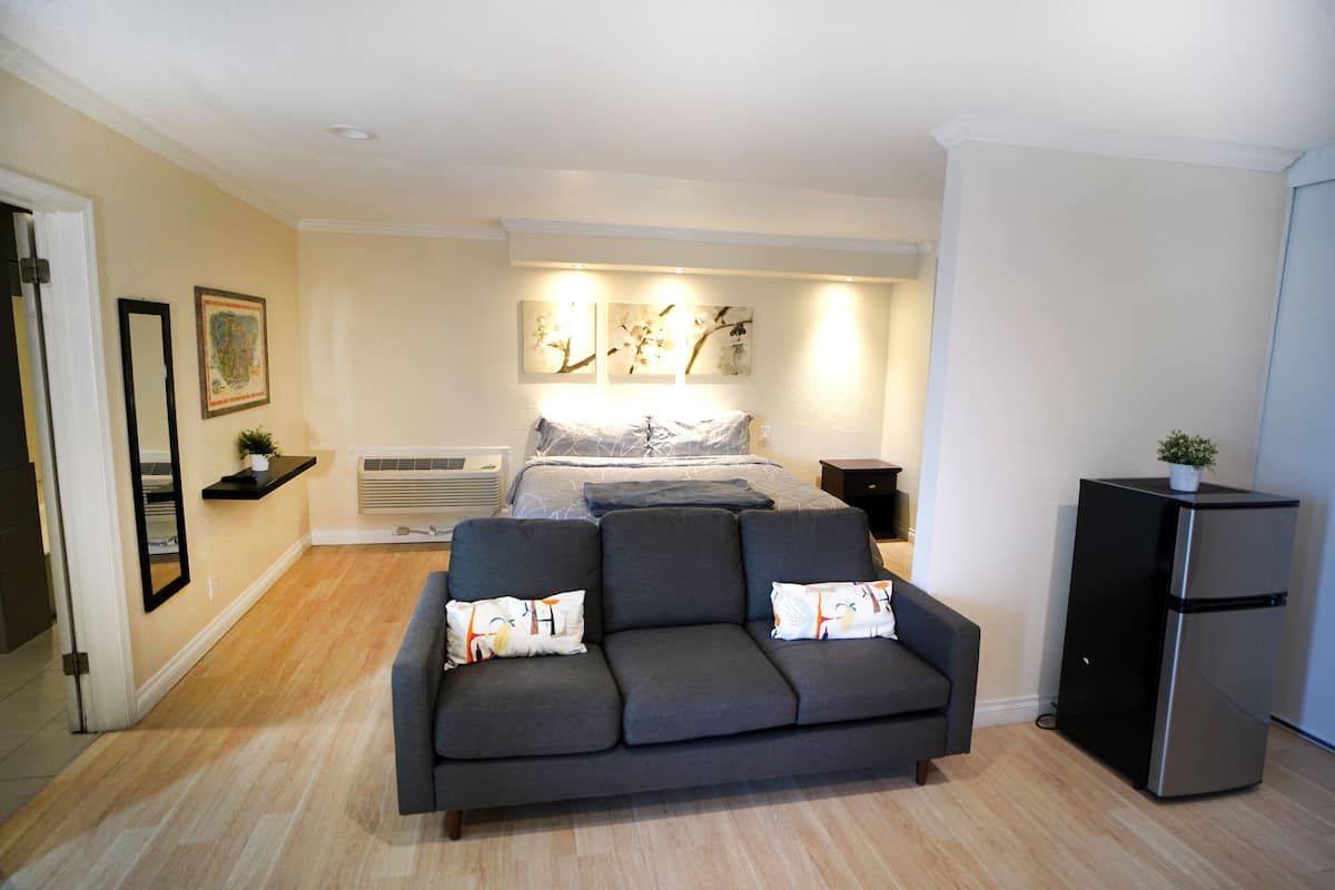 A compact bedroom with a bed, a grey sofa, a small black cabinet, and a wall-mounted air conditioning unit.