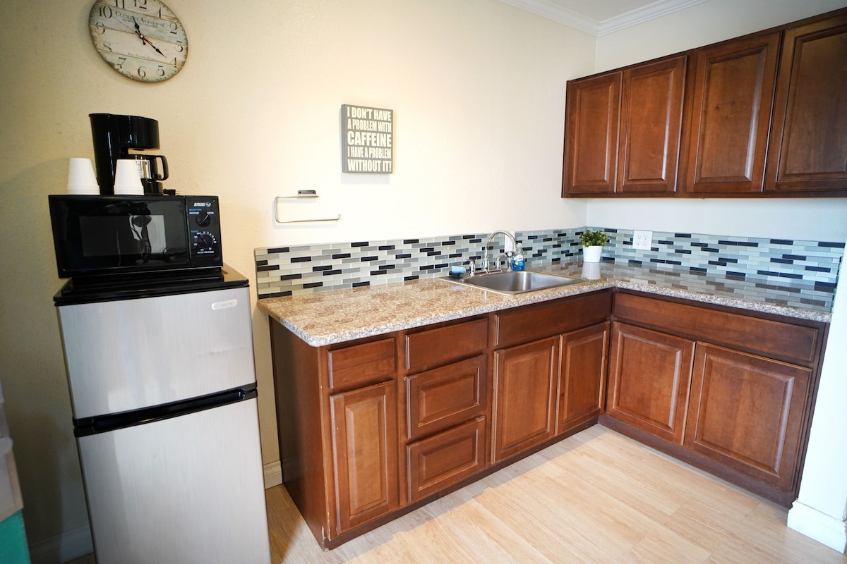 A kitchen with wooden cabinets, a microwave, a refrigerator, a coffee maker, a clock, and a tiled backsplash.