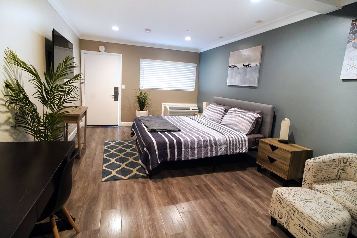 A modern bedroom with a large bed, a desk, and a chair. There is a window with blinds, a plant, and a painting on the wall.