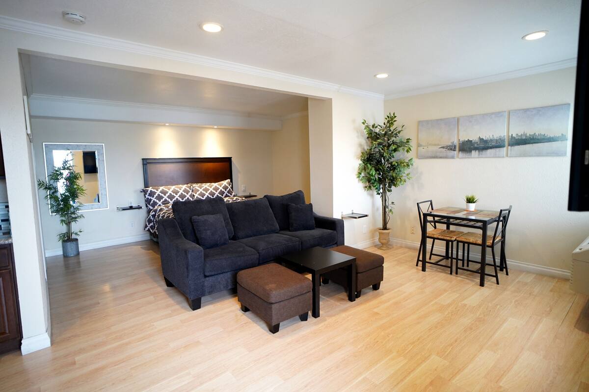 A modern living room with a dark-colored sofa, a matching ottoman, and a wooden coffee table. There is a dining area with a table and chairs, and a large potted plant.