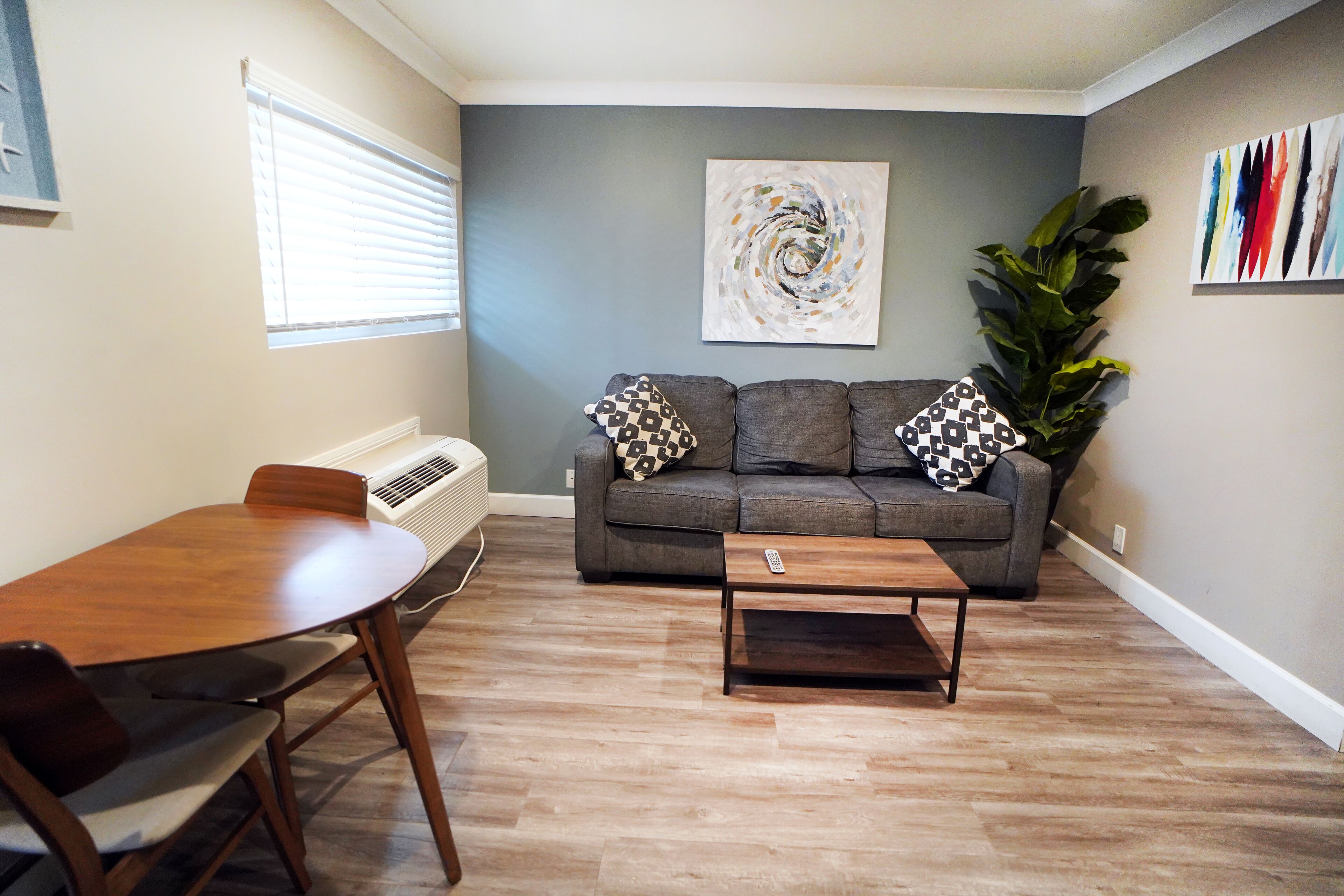 A living room with a gray sofa, two patterned pillows, a wooden coffee table, a dining table with chairs, a large plant, and a wall-mounted abstract painting.